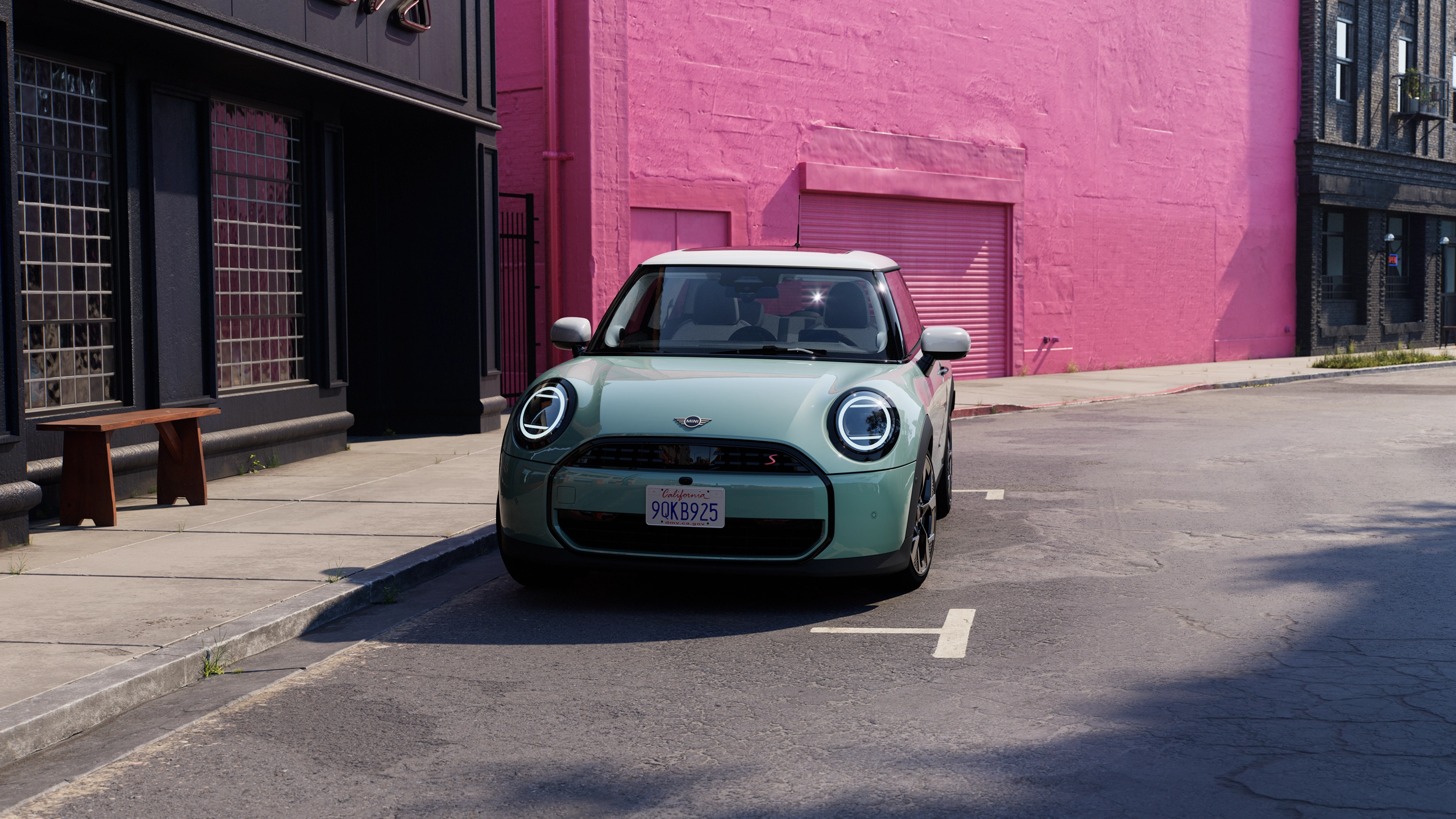 Pohled zepředu na parkující MINI Cooper v zelené barvě Ocean Wave Green s bílou střechou na silnici s růžovou zdí v pozadí