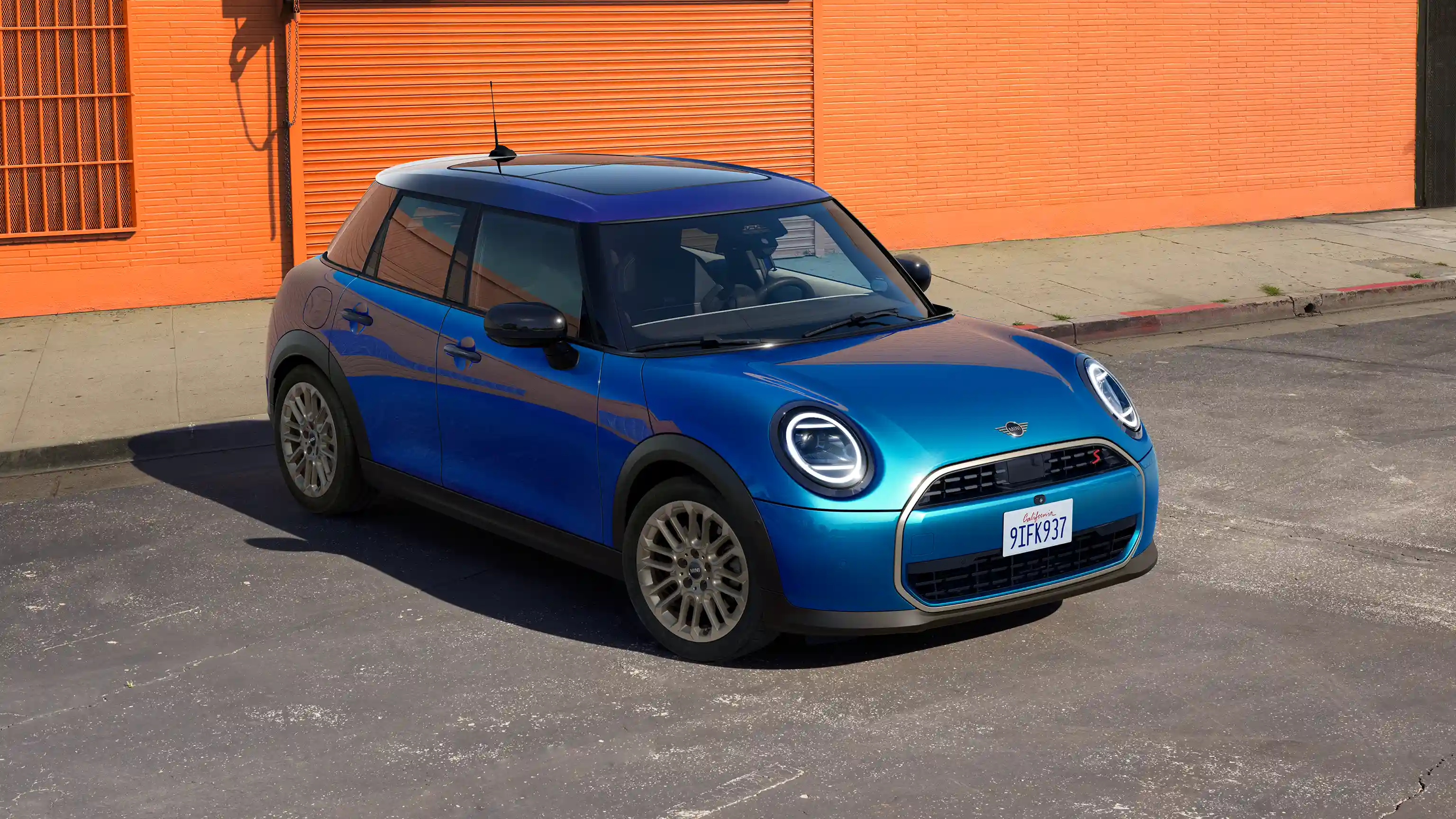 Pohled zepředu na parkující 5dveřové MINI Cooper v modré barvě Icy Sunshine Blue s modrou střechou Multitone Blue na silnici s oranžovou zdí v pozadí.