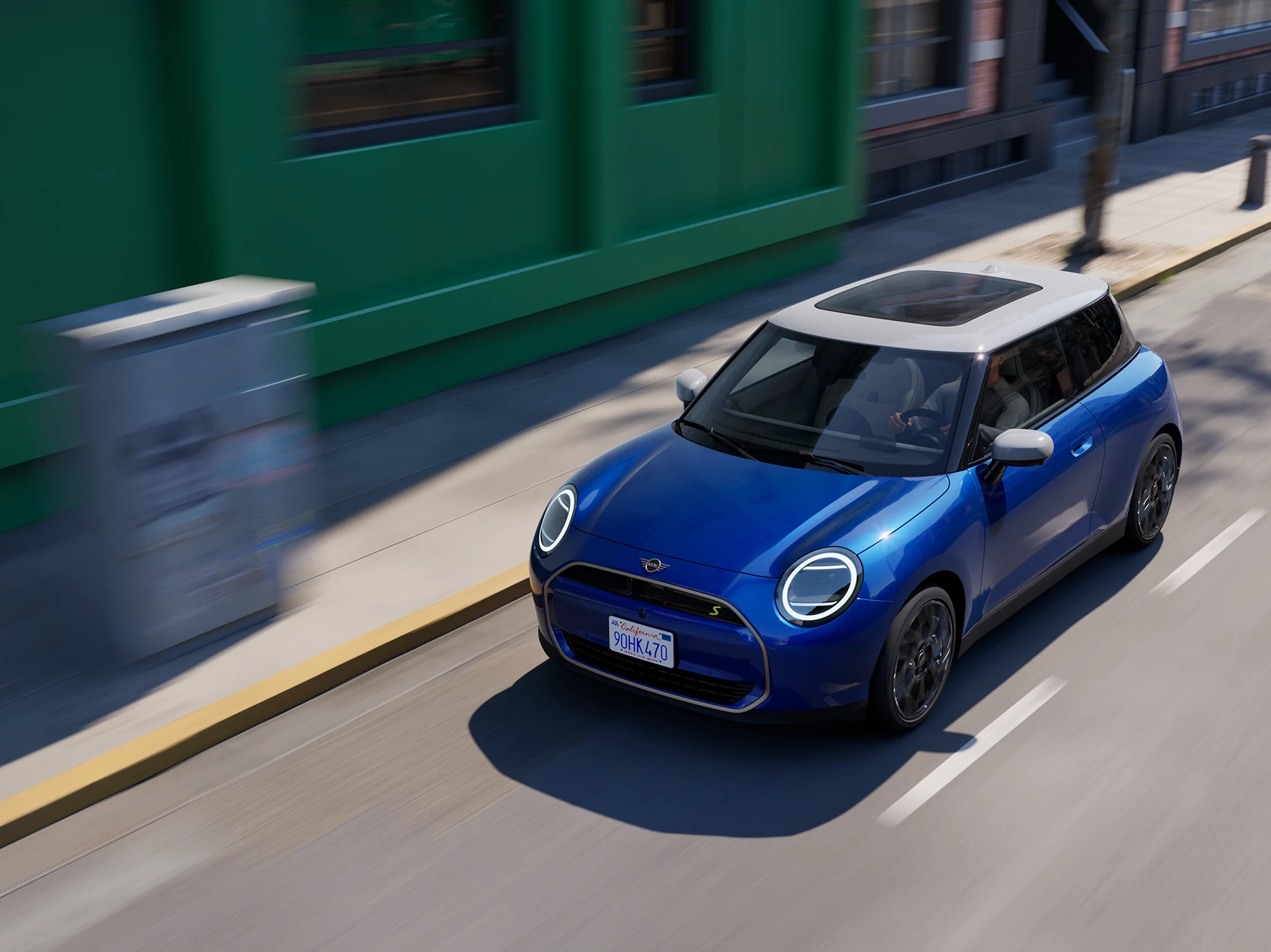 Pohled shora na elektrické MINI Cooper v modré barvě Blazing Blue s bílou střechou jedoucí po ulici. 