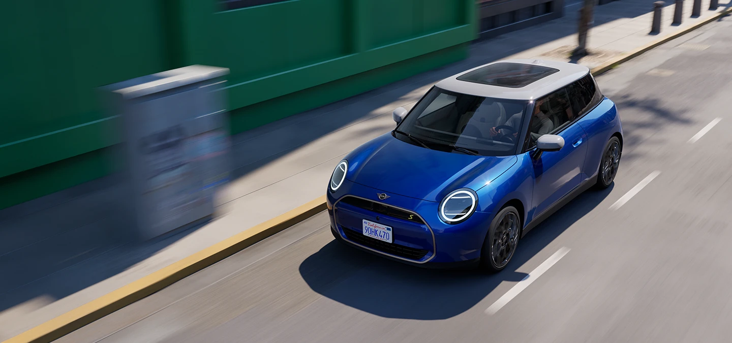Pohled shora na elektrické MINI Cooper v modré barvě Blazing Blue s bílou střechou jedoucí po ulici. 