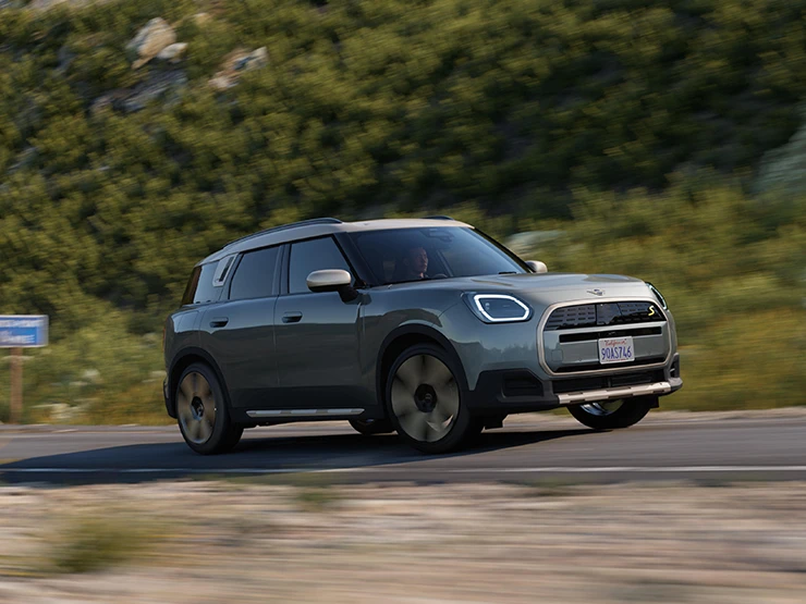 Boční pohled na jedoucí elektrické MINI Countryman v zelené barvě Smokey Green se stříbrnou střechou Vibrant Silver v terénu.