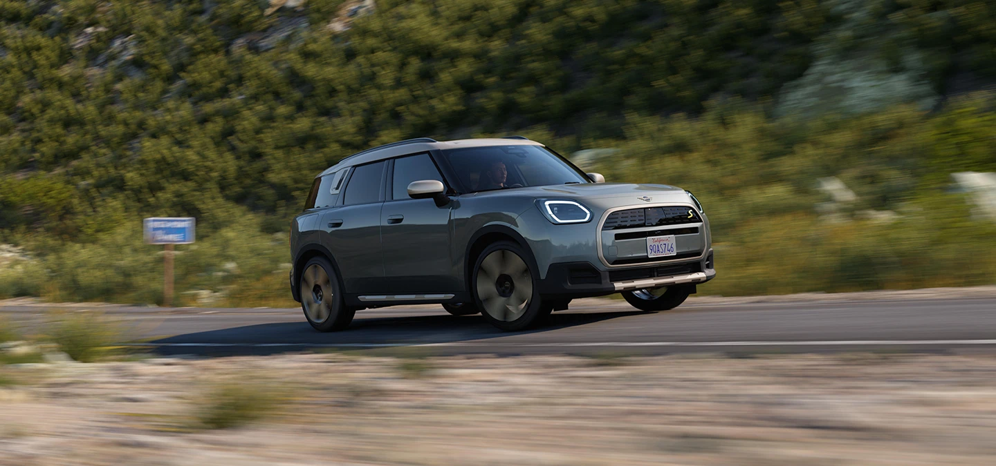 Boční pohled na jedoucí elektrické MINI Countryman v zelené barvě Smokey Green se stříbrnou střechou Vibrant Silver v terénu.