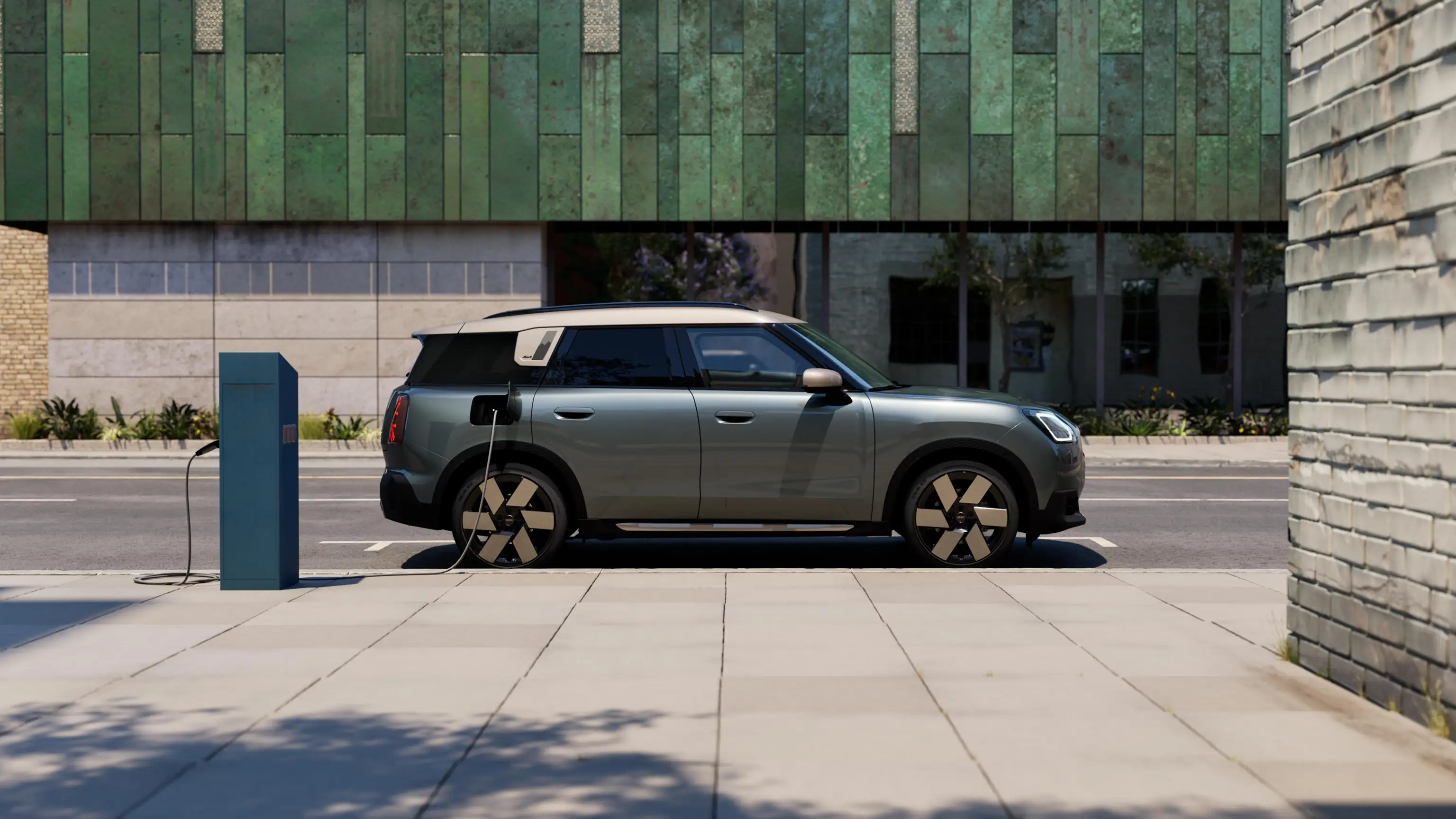 Boční pohled na nabíjející se elektrické MINI Countryman v zelené barvě Smokey Green se stříbrnou střechou Vibrant Silver na ulici