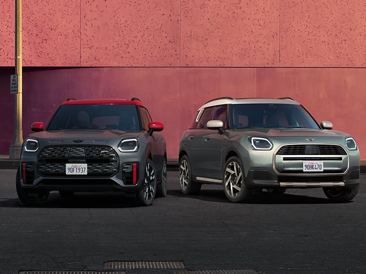 Pohled zepředu na MINI Countryman JCW v šedé barvě Legend Grey a s červenou střechou a na MINI Countryman v zelené barvě Smokey Green a se stříbrnou střechou Vibrant Silver stojící vedle sebe.