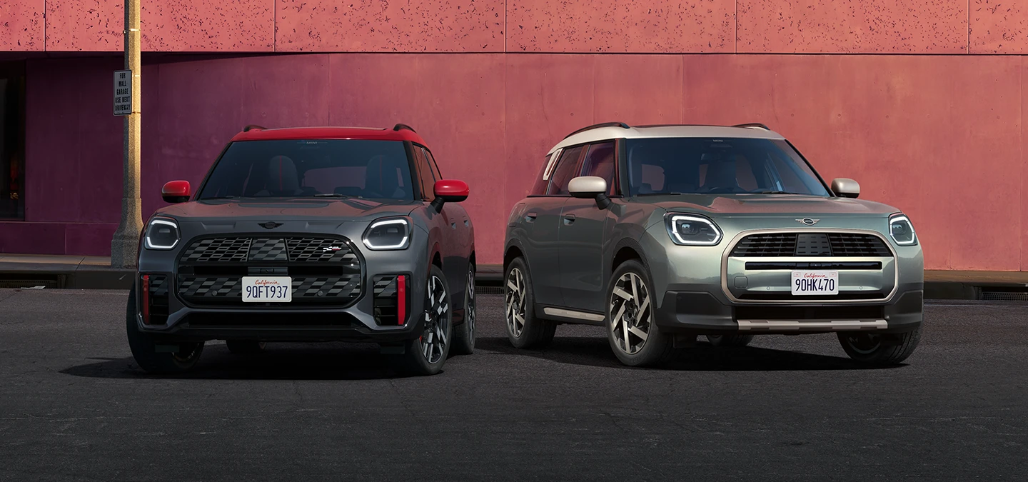 Pohled zepředu na MINI Countryman JCW v šedé barvě Legend Grey a s červenou střechou a na MINI Countryman v zelené barvě Smokey Green a se stříbrnou střechou Vibrant Silver stojící vedle sebe.