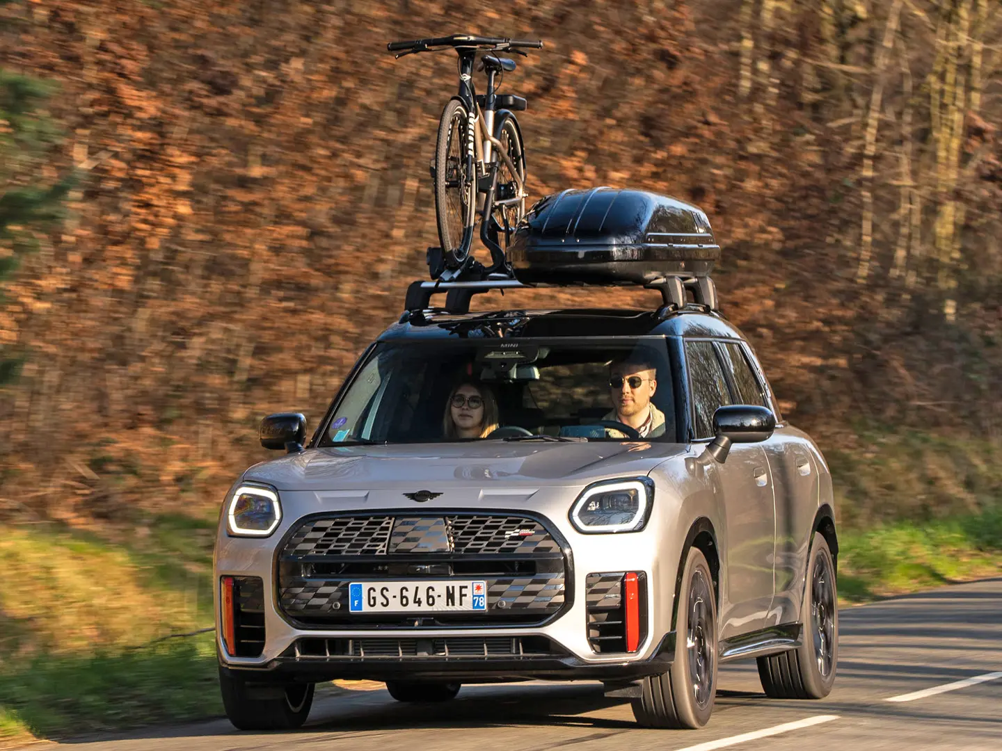 MINI Countryman Cooper ve stříbrné barvě na horské silnici s MINI Střešním nosičem, MINI Střešním boxem, MINI Nosičem kol s jedním namontovaným kolem a wingletem MINI John Cooper Works – pohled zepředu. 
