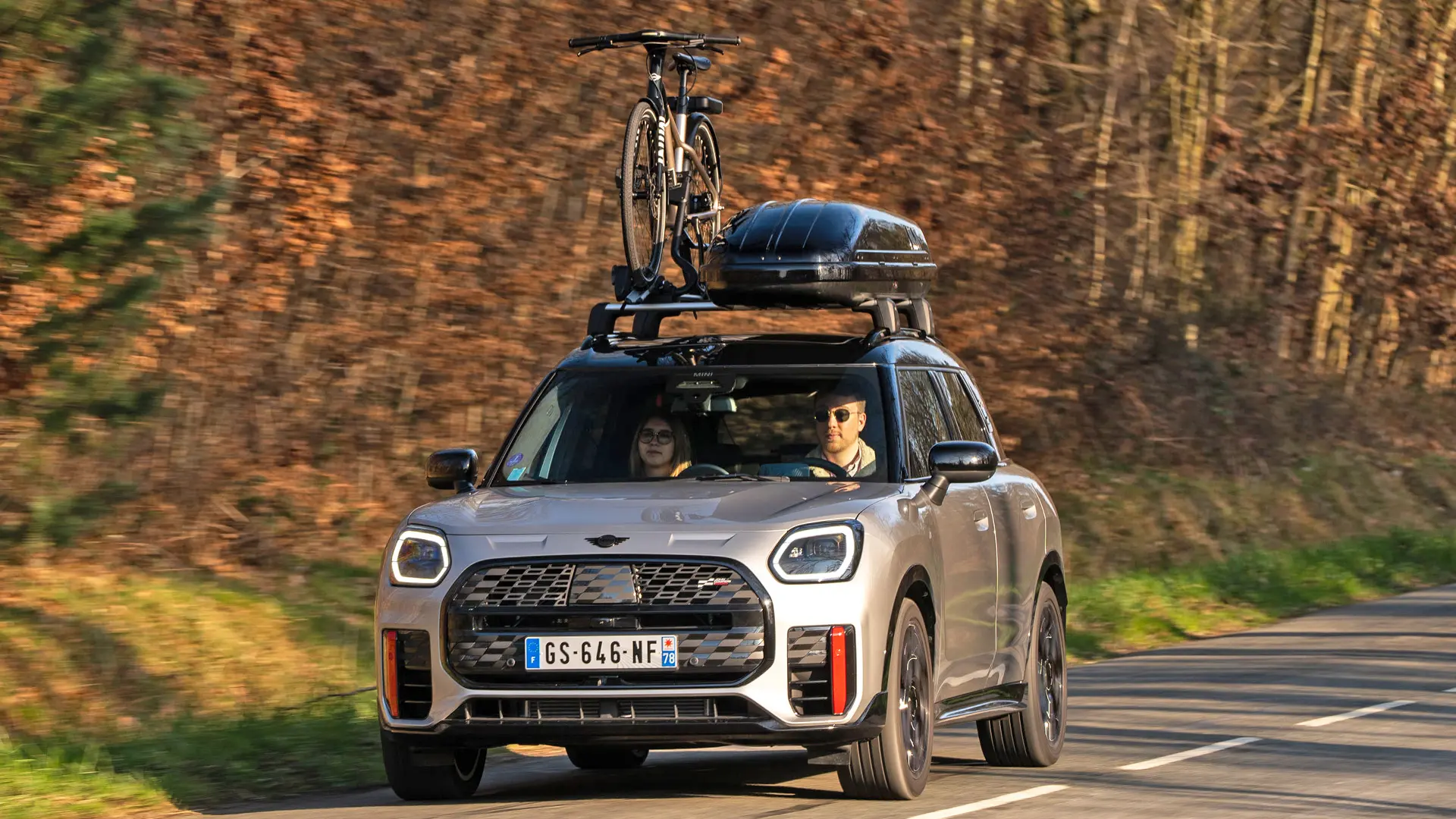 MINI Countryman Cooper ve stříbrné barvě na horské silnici s MINI Střešním nosičem, MINI Střešním boxem, MINI Nosičem kol s jedním namontovaným kolem a wingletem MINI John Cooper Works – pohled zepředu. 