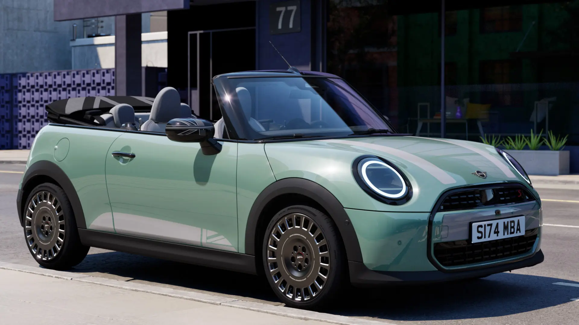 Pohled zepředu na MINI Cabrio v zelené barvě Ocean Wave Green s polepy MINI Heritage na kapotě v bílé barvě. 