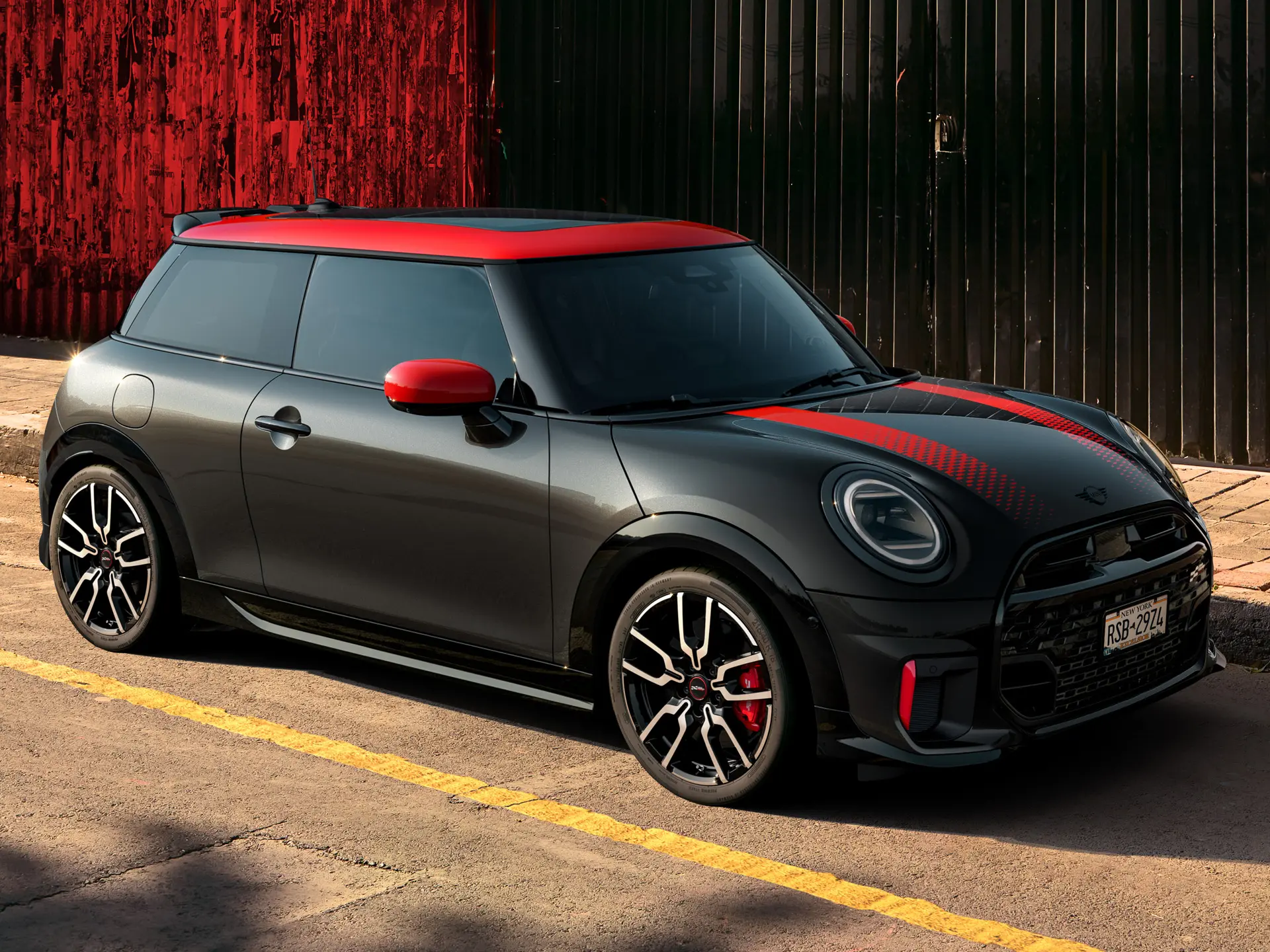 Třídveřové MINI John Cooper Works v černé barvě Midnight Black se střechou a kryty zrcátek v červené barvě Chili Red a červenými sportovními pruhy, pohled z boku, zaparkované na ulici.