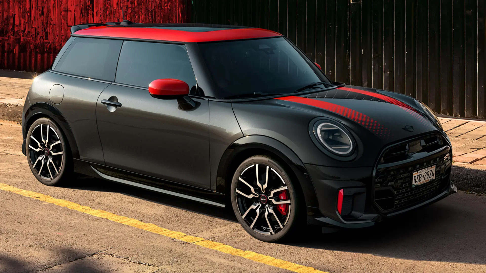 Třídveřové MINI John Cooper Works v černé barvě Midnight Black se střechou a kryty zrcátek v červené barvě Chili Red a červenými sportovními pruhy, pohled z boku, zaparkované na ulici.