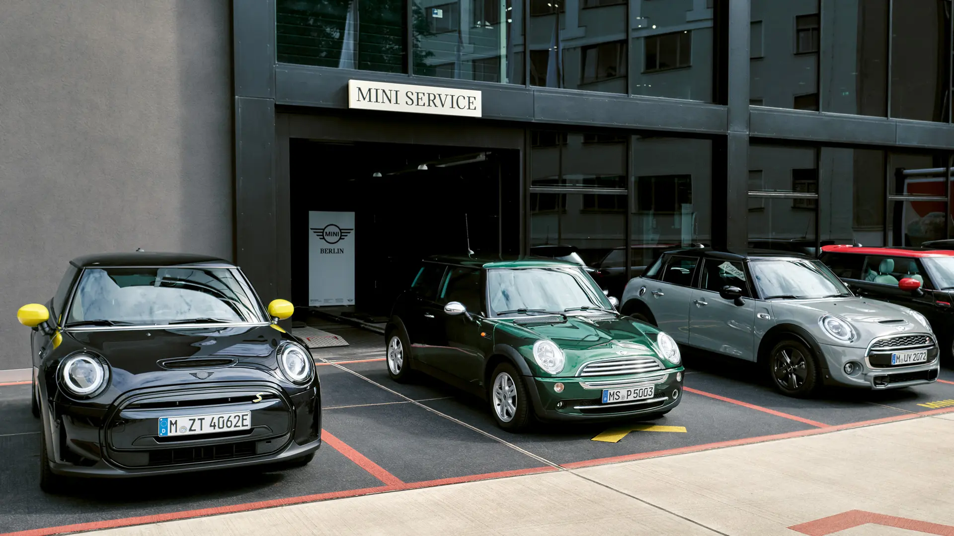 Elektrické MINI Cooper S v černé barvě stojí před MINI Centrem vedle třídveřového MINI Cooper v zelené barvě, pětidveřového MINI ve stříbrné barvě a černého MINI JCW, pohled zepředu. 