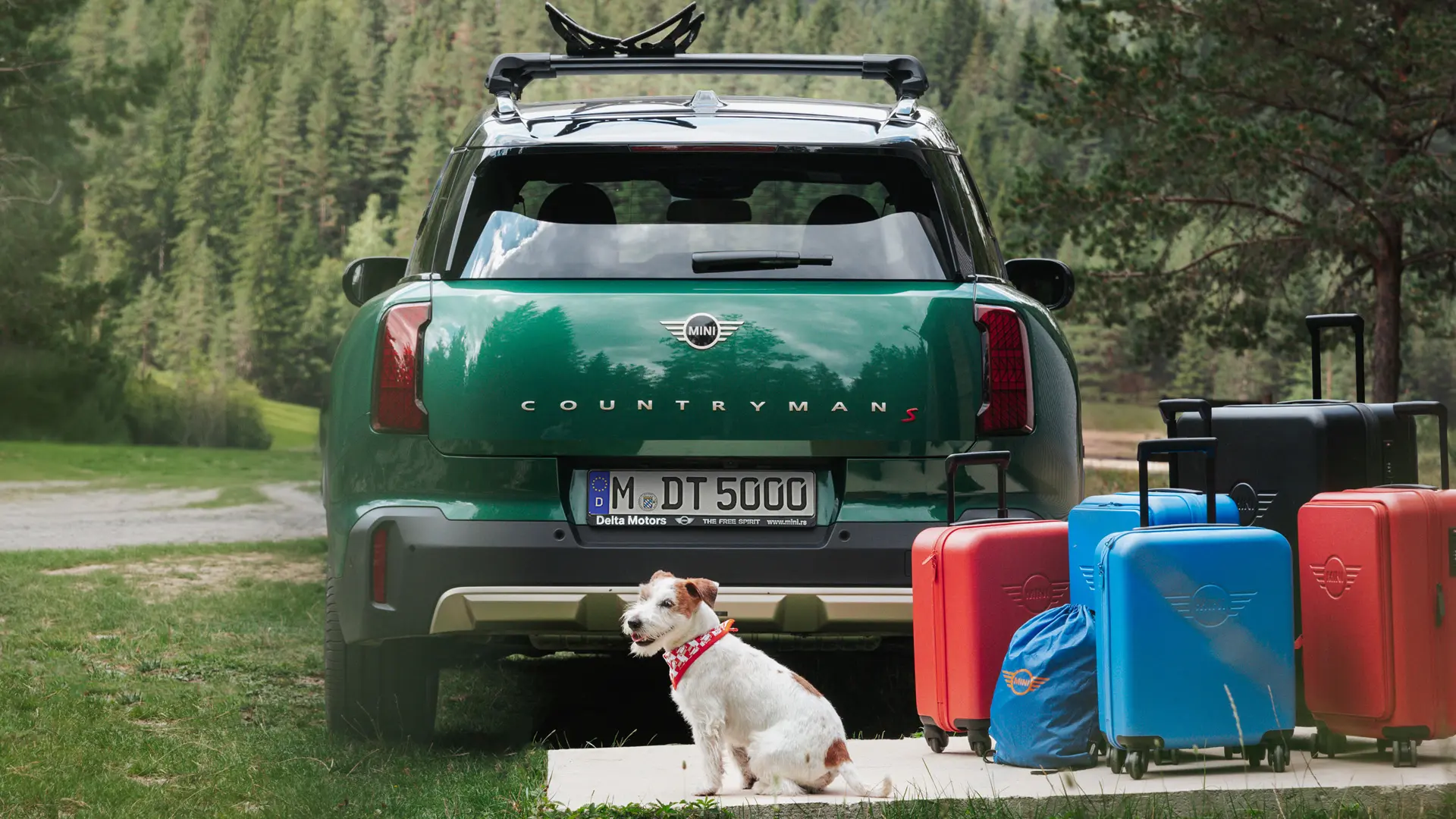 MINI Countryman v zelené barvě s různými zavazadly dostupnými v obchodě MINI Lifestyle stojící za vozidlem společně se psem Jackem Russellem. 