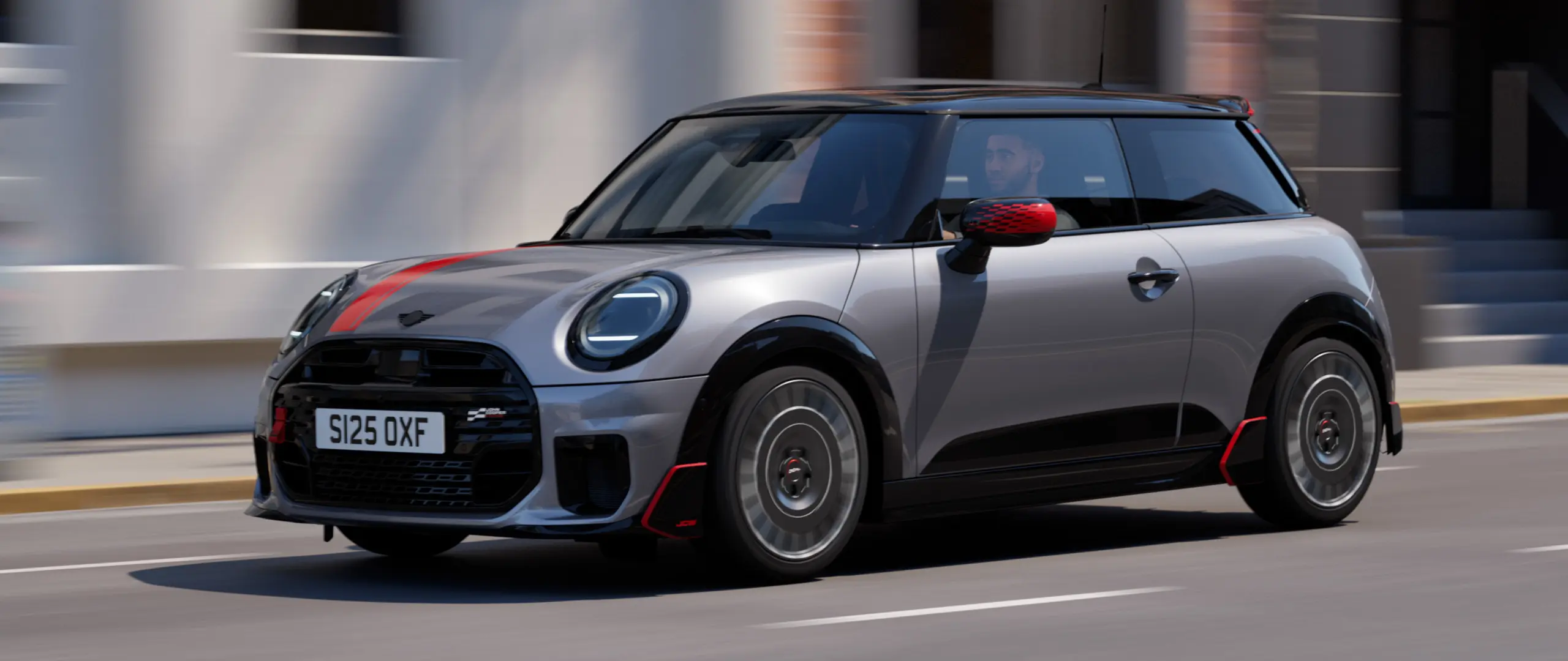 3dveřové MINI Cooper ve stříbrné barvě Melting Silver s John Cooper Works kryty zpětných zrcátek, červenými polepy na kapotě Chili Red, střešním spoilerem a černým wingletem jedoucí po městské ulici – tříčtvrteční pohled zepředu.